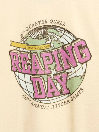 A close-up of a beige garment featuring a large graphic print for the 50th Annual Hunger Games, titled 'Reaping Day,' with a globe illustration and pink lettering.