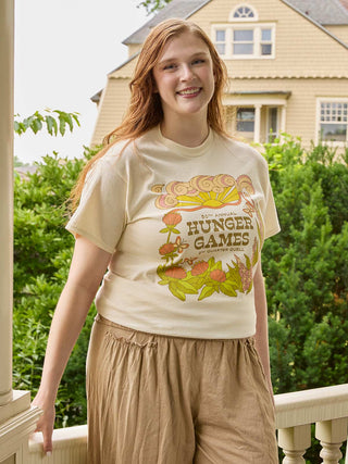 A smiling young woman stands on a balcony wearing a cream-colored graphic t-shirt with a retro 'Hunger Games' design and tan drawstring pants, with a house and green bushes in the background.