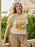 A smiling young woman stands on a balcony wearing a cream-colored graphic t-shirt with a retro 'Hunger Games' design and tan drawstring pants, with a house and green bushes in the background.