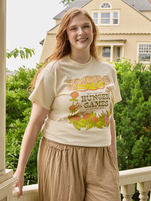 A smiling young woman stands on a balcony wearing a cream-colored graphic t-shirt with a retro 'Hunger Games' design and tan drawstring pants, with a house and green bushes in the background.