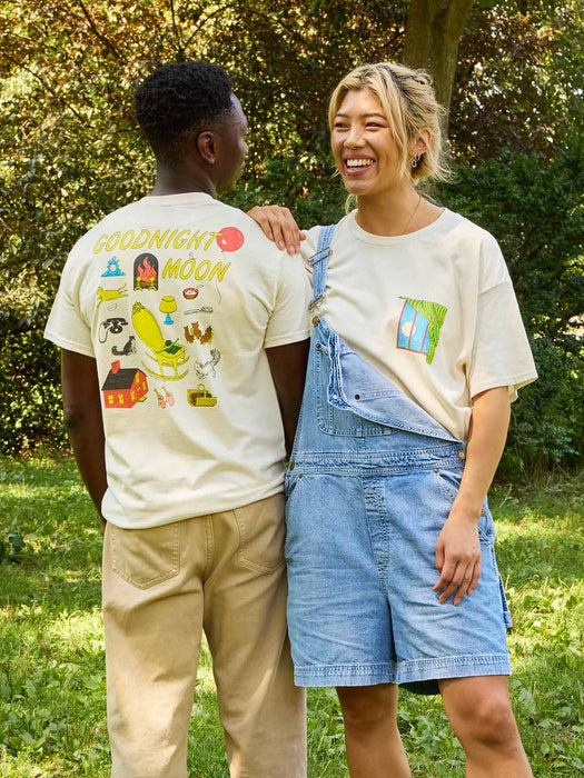 Two models standing outdoors in a grassy park setting, showcasing a matching cream-colored graphic t-shirt. The back of the shirt features colorful illustrations with the text 'GOODNIGHT MOON', while the front shows a small colorful graphic on the chest.