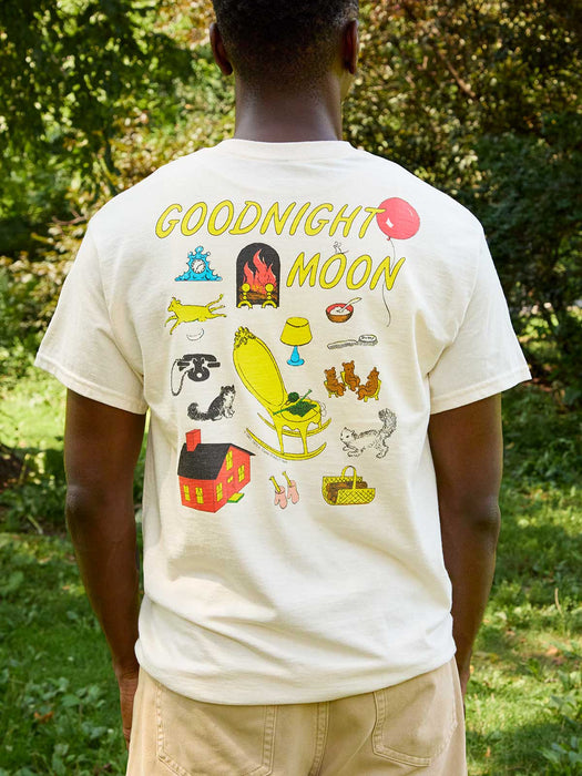 A man viewed from behind wearing a cream-colored t-shirt with a colorful graphic print on the back that reads 'GOODNIGHT MOON' and features various whimsical illustrations, set against a background of green trees and grass.