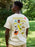 A man wearing a cream-colored t-shirt with a colorful graphic print on the back, standing outdoors in a grassy, wooded area. The graphic features the text 'GOODNIGHT MOON' in yellow above various whimsical illustrations.