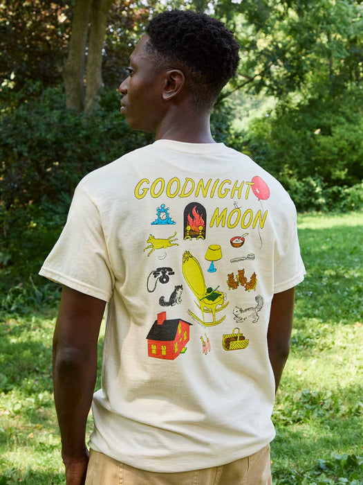 A man wearing a cream-colored t-shirt with a colorful graphic print on the back, standing outdoors in a grassy, wooded area. The graphic features the text 'GOODNIGHT MOON' in yellow above various whimsical illustrations.