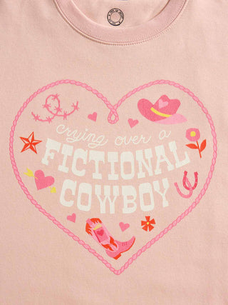 A close-up of a light pink t-shirt featuring a heart-shaped graphic with the words 'crying over a FICTIONAL COWBOY' in white and pink, surrounded by cowboy hat, horseshoe, star, and flower illustrations.