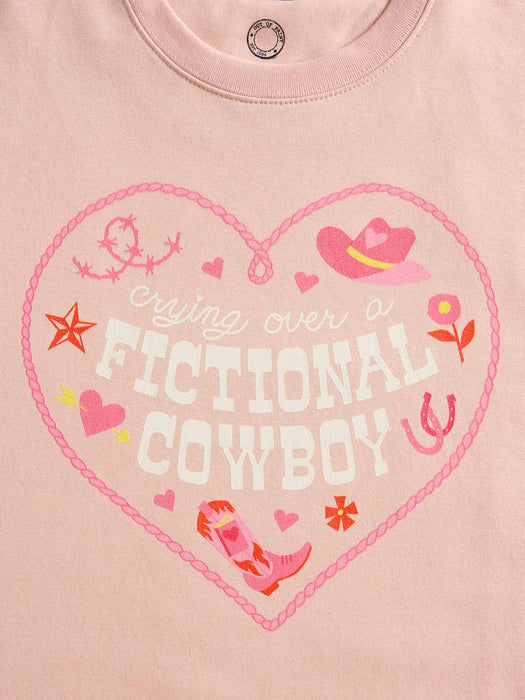 A close-up of a light pink t-shirt featuring a heart-shaped graphic with the words 'crying over a FICTIONAL COWBOY' in white and pink, surrounded by cowboy hat, horseshoe, star, and flower illustrations.