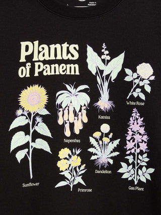 A close-up of a black t-shirt featuring a graphic print titled 'Plants of Panem'. The design displays various illustrated flowers and plants with their names printed below them.