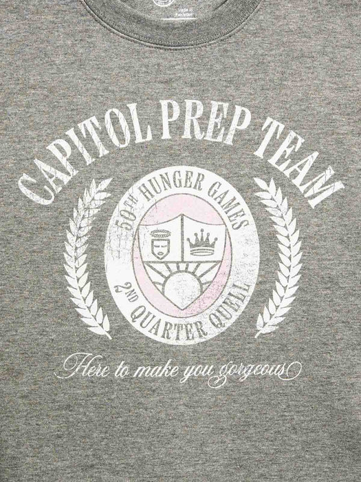 Close-up of a heather gray crewneck t-shirt featuring a distressed white graphic print that reads 'CAPITOL PREP TEAM' with a central Hunger Games emblem and the phrase 'Here to make you gorgeous'.