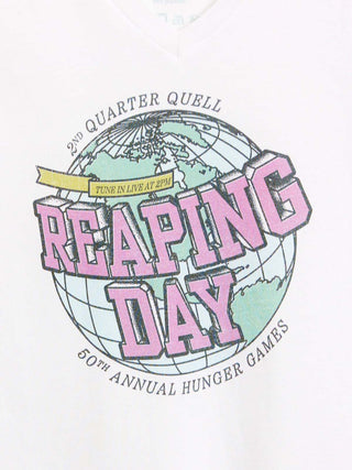 A close-up of a white garment featuring a graphic print for the 50th Annual Hunger Games 'Reaping Day' event. The design includes a globe illustration with pink 3D text reading 'REAPING DAY' and smaller black text reading '2ND QUARTER QUELL' and '50TH ANNUAL HUNGER GAMES'.