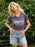 A woman wearing a heather navy blue graphic t-shirt with 'ASTRONAUTICALS' printed in pink and white, paired with light-wash denim shorts, standing in front of a green leafy background.