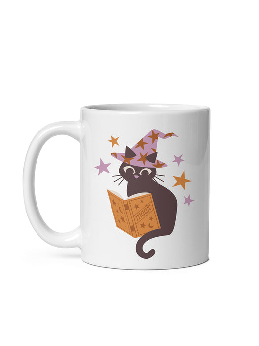 A white ceramic mug featuring a cartoon illustration of a black cat wearing a purple witch hat and reading an orange magic book, surrounded by small stars.