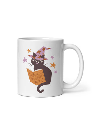 A white ceramic mug featuring a cartoon illustration of a black cat wearing a purple witch hat and reading an orange magic book, surrounded by small stars.