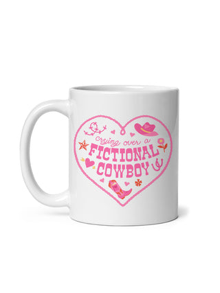 A white ceramic mug with a pink heart-shaped graphic that reads 'crying over a FICTIONAL COWBOY' in stylized lettering, decorated with cowboy hat, boots, stars, and small pink accents, shown on a white background.