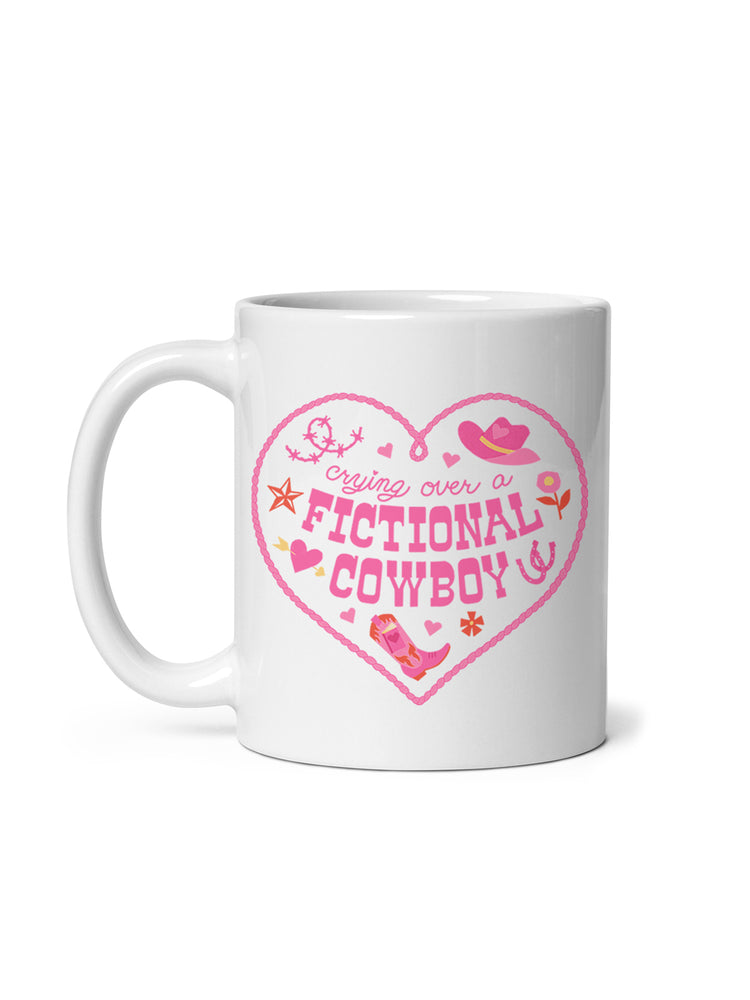 A white ceramic mug with a pink heart-shaped graphic that reads 'crying over a FICTIONAL COWBOY' in stylized lettering, decorated with cowboy hat, boots, stars, and small pink accents, shown on a white background.