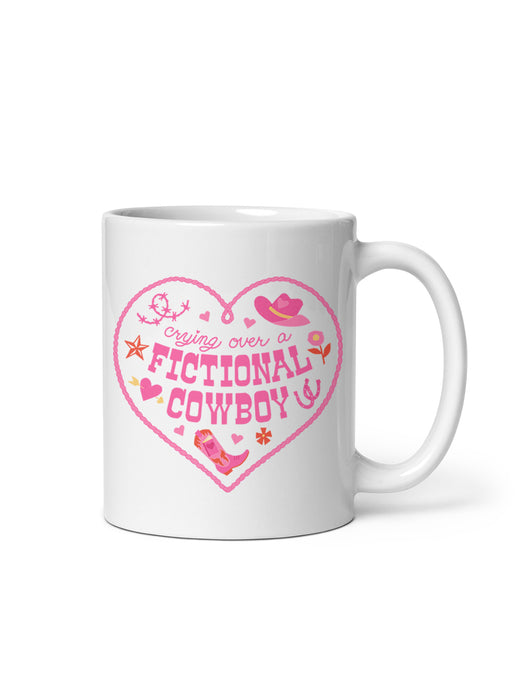 A white ceramic mug with a pink graphic on the front that reads 'Crying over a FICTIONAL COWBOY' inside a heart outline, with small cowboy boot, hat, and star details, shown on a white background.