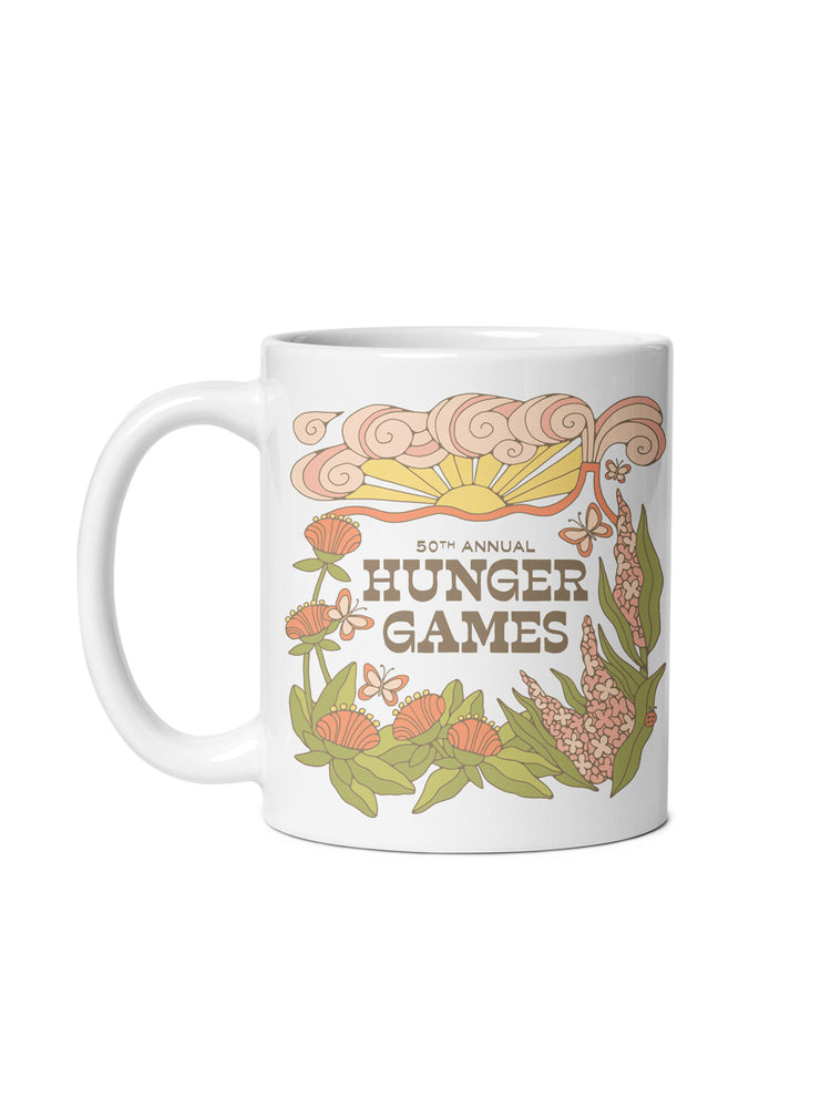 A white ceramic mug featuring a retro-style graphic design for the 50th Annual Hunger Games, with stylized flowers, leaves, and a sun in muted orange, green, and brown tones.