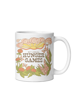 A white ceramic mug featuring a retro-style illustrated design for the 50th Annual Hunger Games, with peach clouds, flowers, butterflies, and green leaves on a white background.