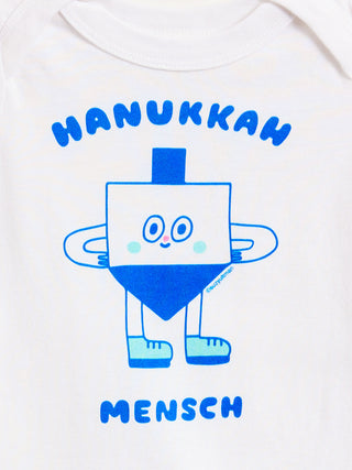 White baby bodysuit featuring a blue graphic of a smiling dreidel character with the text 'HANUKKAH' above and 'MENSCH' below.