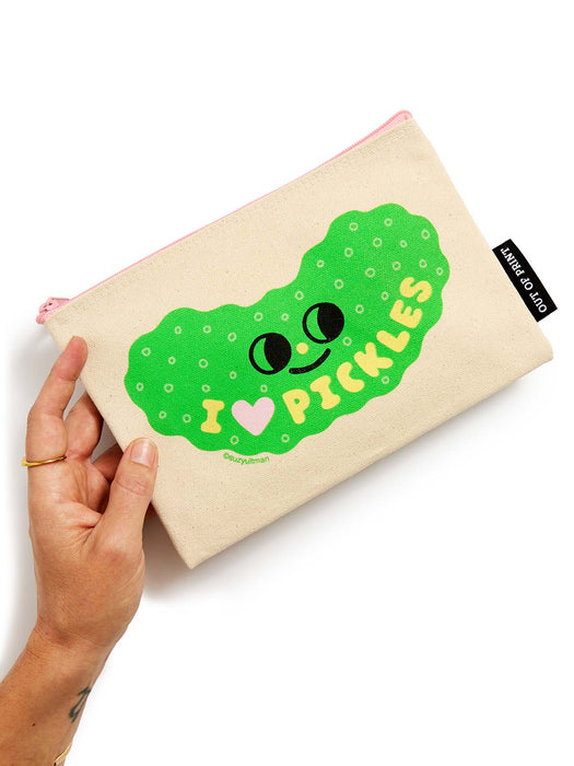 A hand holding a beige canvas pouch with a green pickle graphic and the text 'I [heart] PICKLES'. The pouch has a pink zipper and a black 'OUT OF PRINT brand tag on the side.