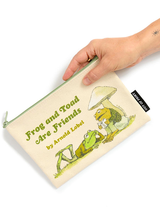 A hand holds a cream-colored canvas pouch featuring the classic 'Frog and Toad Are Friends' illustration and text by Arnold Lobel. The pouch has a light green zipper and a black fabric brand tag on the right side that reads 'OUT OF PRINT'