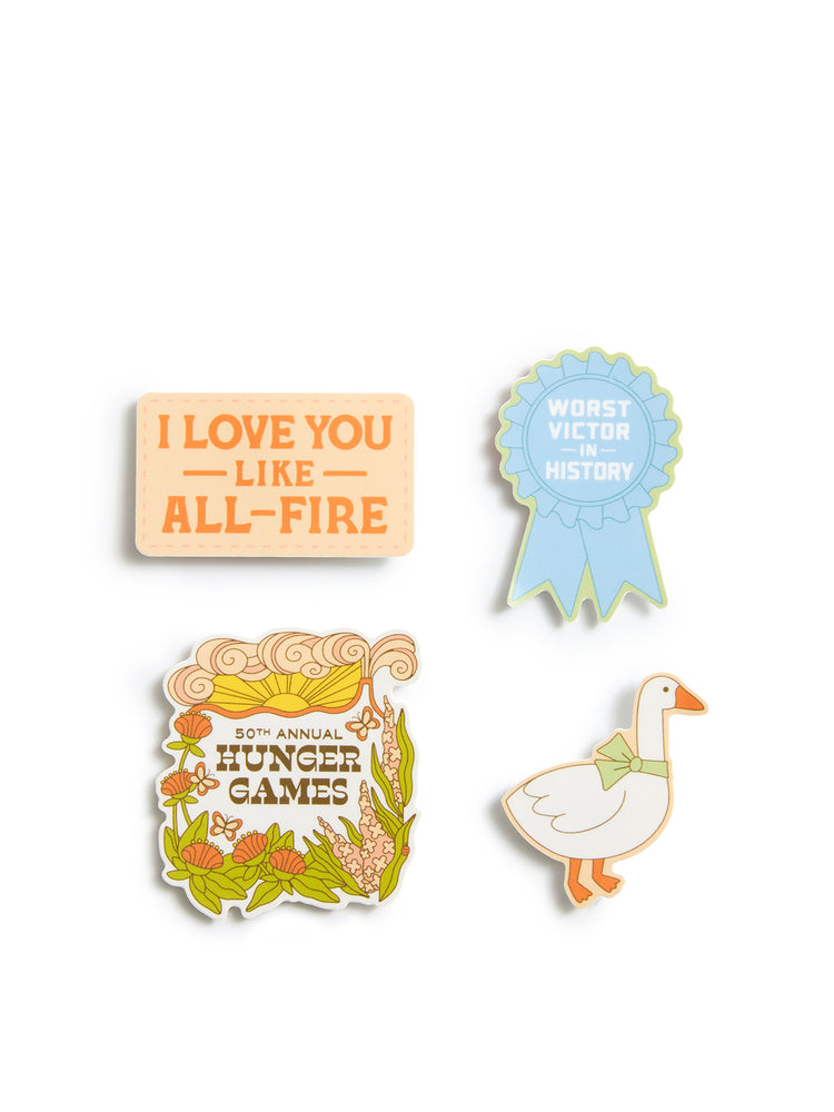 Four stickers arranged on a plain white background: a rectangular sticker reading 'I LOVE YOU LIKE ALL-FIRE,' a light blue ribbon reading 'WORST VICTOR IN HISTORY,' a floral sticker reading '50th Annual Hunger Games,' and a white goose sticker with orange and green details.