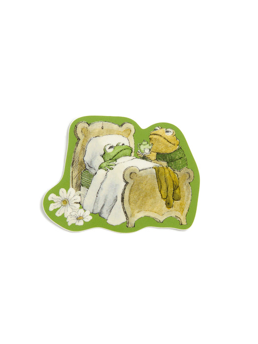 A die-cut sticker featuring a whimsical illustration of a green frog in bed with a white blanket set against a light green background with white daisy flowers in the lower left corner and Toad holding a tea kettle.