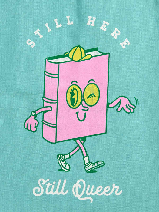 A graphic print on a tote bag featuring a pink cartoon book character with yellow eyes and limbs, with the text 'STILL HERE' above and 'Still Queer' below.