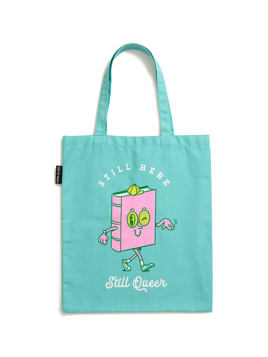 A light teal canvas tote bag featuring a cute pink book character with a smiling face, the words 'STILL HERE' above it, and 'Still Queer' written below in white script.