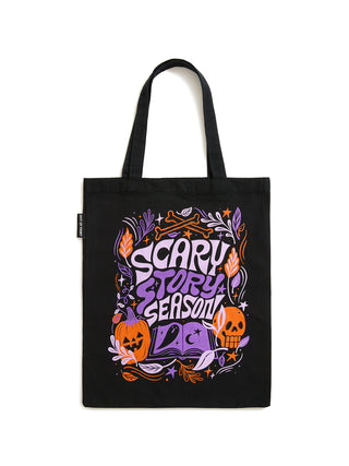 A black canvas tote bag with a colorful Halloween-themed graphic that reads 'SCARY STORY SEASON' in purple and orange lettering, featuring pumpkins, a skull, and decorative foliage, shown against a plain white background.