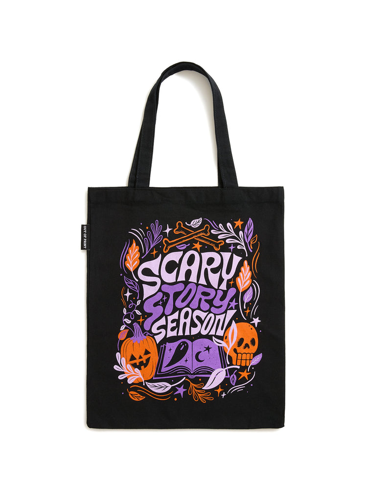 A black canvas tote bag with a colorful Halloween-themed graphic that reads 'SCARY STORY SEASON' in purple and orange lettering, featuring pumpkins, a skull, and decorative foliage, shown against a plain white background.