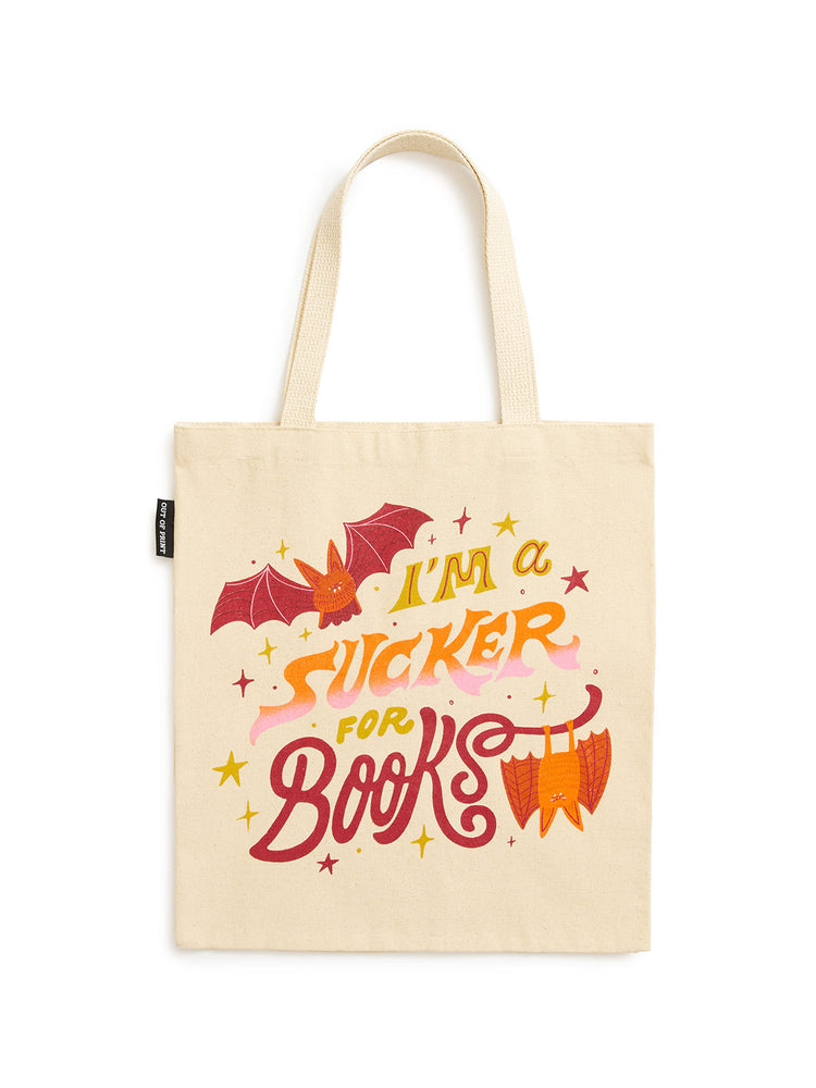 A cream-colored canvas tote bag with a colorful graphic print that reads 'I'M A SUCKER FOR BOOKS' in stylized red, orange, and yellow lettering, accompanied by illustrations of a bat and a book.