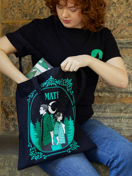Person holding a tote bag with a design of two people and the word 'MATE' on a stone wall background