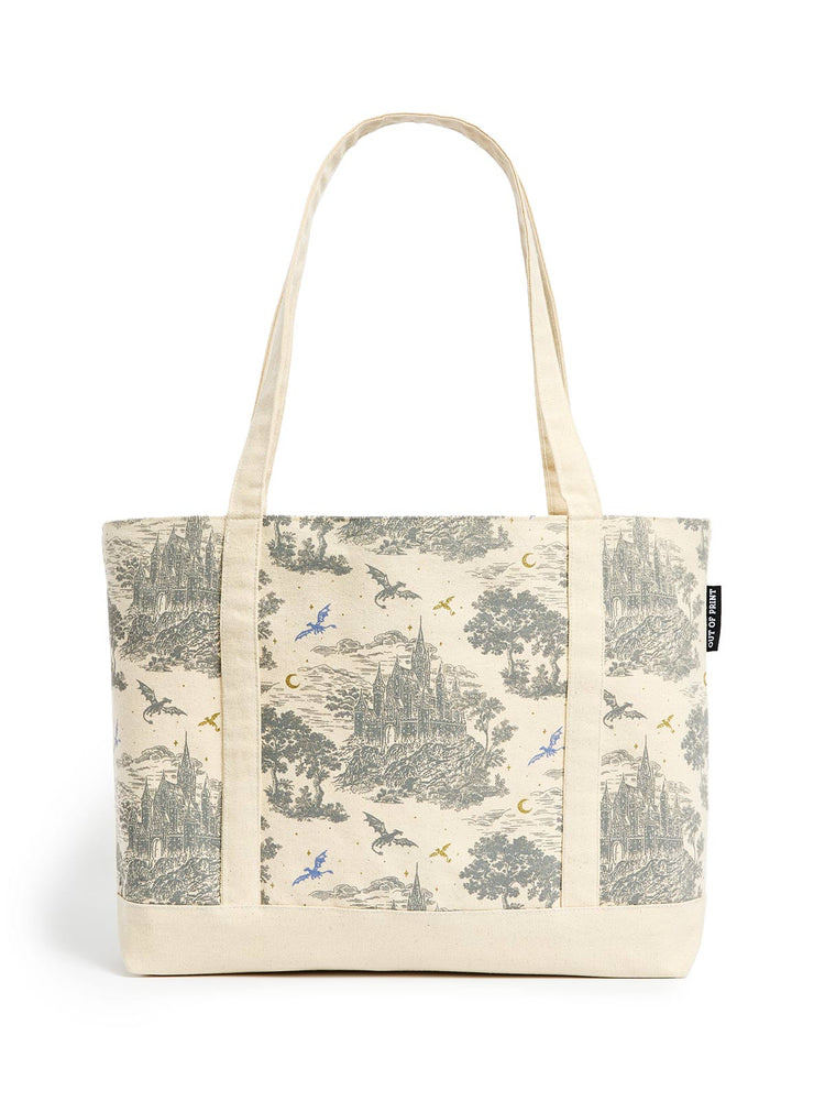 A canvas tote bag with a cream-colored base and a detailed toile-style print featuring castles, trees, and flying dragons in muted gray, blue, and gold tones. The bag has two long cream handles and a small black brand tag on the right side.