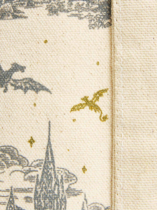 Close-up of a woven textile with an off-white background, featuring a gray printed topper-style illustration of a spire, clouds, and a dragon, plus small metallic gold star and dragon accents. A vertical stitched seam runs along the right side.