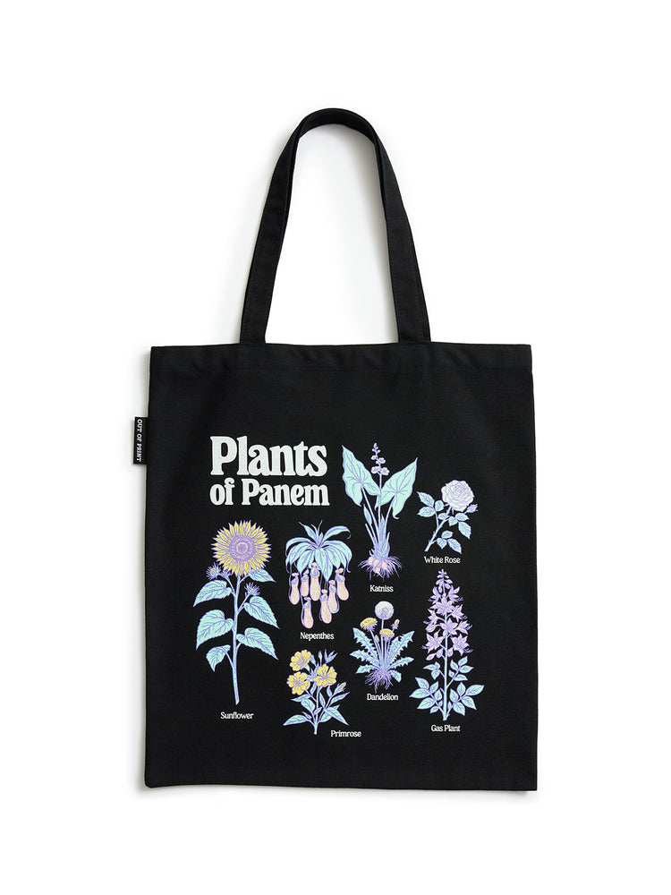 A black canvas tote bag with two shoulder straps, featuring a graphic print titled 'Plants of Panem' with various illustrated plants in pastel colors and small labels, shown against a plain white background.