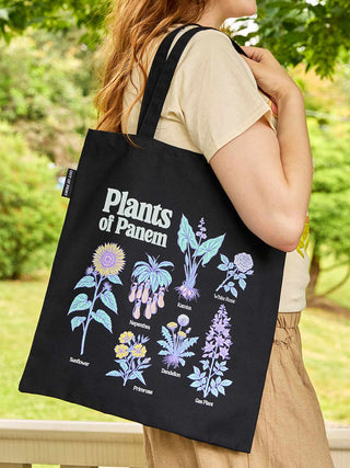 A person outdoors carrying a black canvas tote bag printed with the words "Plants of Panem" and several illustrated plants in pastel colors. The background shows blurred green foliage and grass.