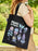 A person outdoors carrying a black canvas tote bag printed with the words "Plants of Panem" and several illustrated plants in pastel colors. The background shows blurred green foliage and grass.