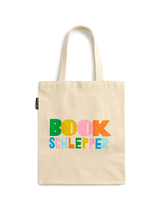 A natural beige canvas tote bag with two shoulder straps, featuring the text 'BOOK SCHLEPPER' printed in colorful, playful letters across the front. A small black fabric tag is visible on the left side seam.