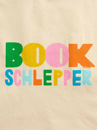 A close-up of a beige fabric surface with the words 'BOOK SCHLEPPER' printed in large, colorful block letters. The letters are filled with green, yellow, pink, orange, blue, and light blue colors.