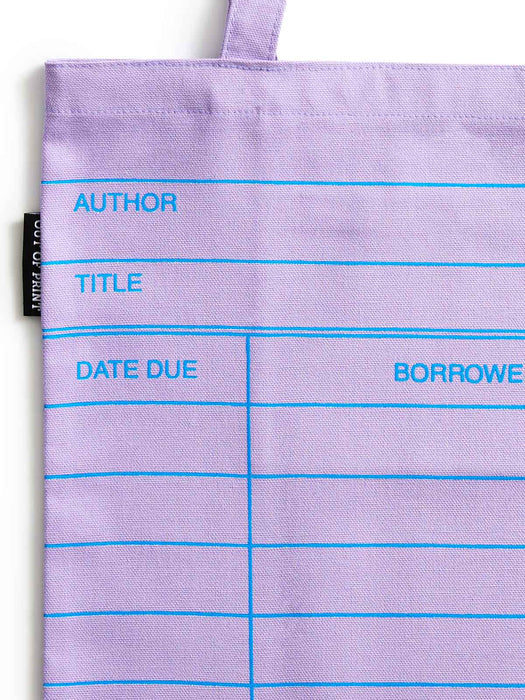 A close-up of a lilac purple canvas tote bag with bright blue printed lines and text fields for author, title, date due, and borrower. A small black fabric tag is visible on the left side.