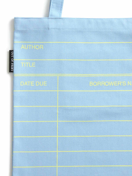 A light cloud blue fabric tote bag with yellow printed lines and text fields for author, title, date due, and borrower's name. A small black fabric tag on the left edge reads 'OUT OF PRINT'.