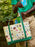 A person outdoors holding a canvas tote bag with green handles and trim, featuring a cream background printed with colorful cartoon illustrations and the words 'GOODNIGHT MOON'.