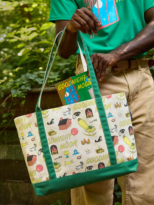 A person outdoors holding a canvas tote bag with green handles and trim, featuring a cream background printed with colorful cartoon illustrations and the words 'GOODNIGHT MOON'.
