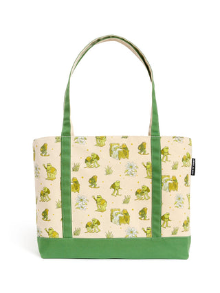 A canvas tote bag with a light beige background printed with green frogs and small white flowers, featuring solid green handles and a green bottom base.