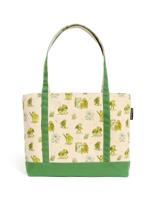 A canvas tote bag with a light beige background printed with green frogs and small white flowers, featuring solid green handles and a green bottom base.