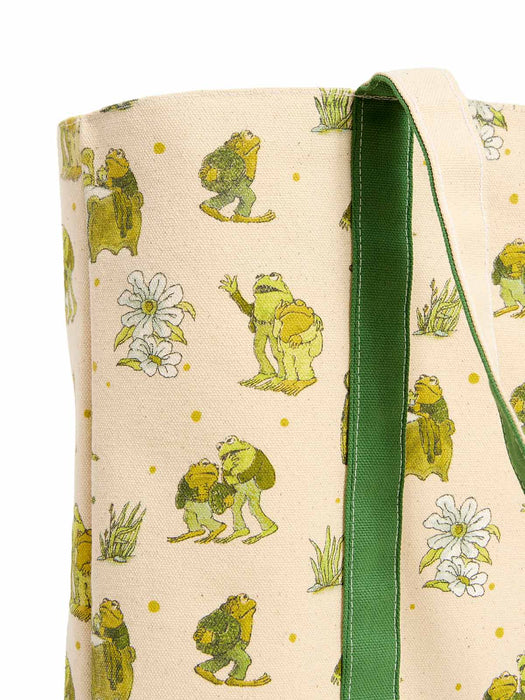 A close-up of a canvas tote bag featuring a whimsical print of green frogs, white flowers, and small yellow dots on a cream background, with a solid green fabric strap.