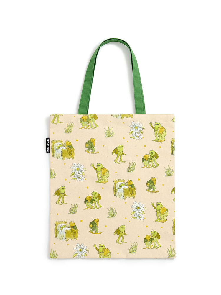 A canvas tote bag with a light beige background printed with a whimsical pattern of green frogs, white flowers, and small yellow dots. The bag has two green fabric handles and a small black brand tag on the left side.