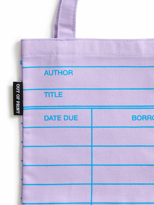 A close-up of a lilac purple tote bag with bright blue printed lines and text fields for author, title, date due, and borrow. A black fabric tag on the left reads OUT OF PRINT.