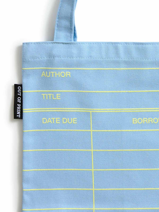 A light cloud blue canvas tote bag with yellow printed library-style fields including AUTHOR, TITLE, DATE DUE, and BORROW, plus a black side tag reading OUT OF PRINT.