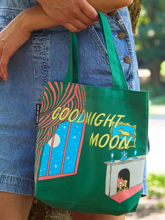 A green canvas mini tote bag with a colorful graphic print of a window showing a full moon and the words 'GOODNIGHT MOON!' in yellow, carried by a person wearing denim shorts outdoors.